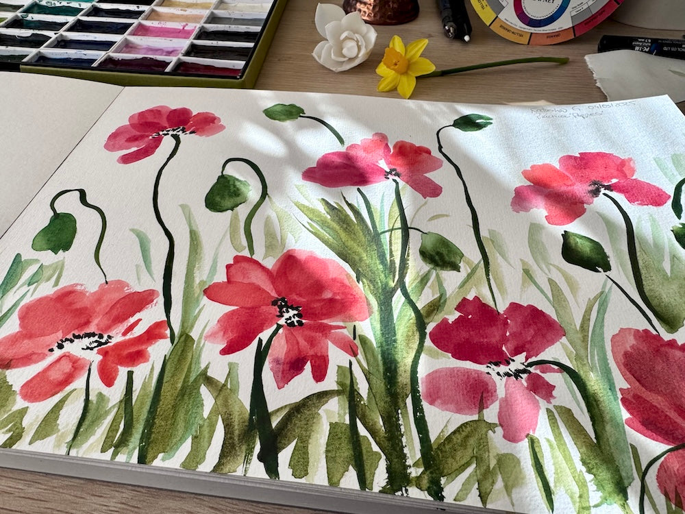 Aquarelle représentant un champ de coquelicots rouges et roses aux tiges élancées, entourés de feuillages verts. En arrière-plan, une palette de peinture, un pinceau et une jonquille posée sur un bureau baigné de lumière.